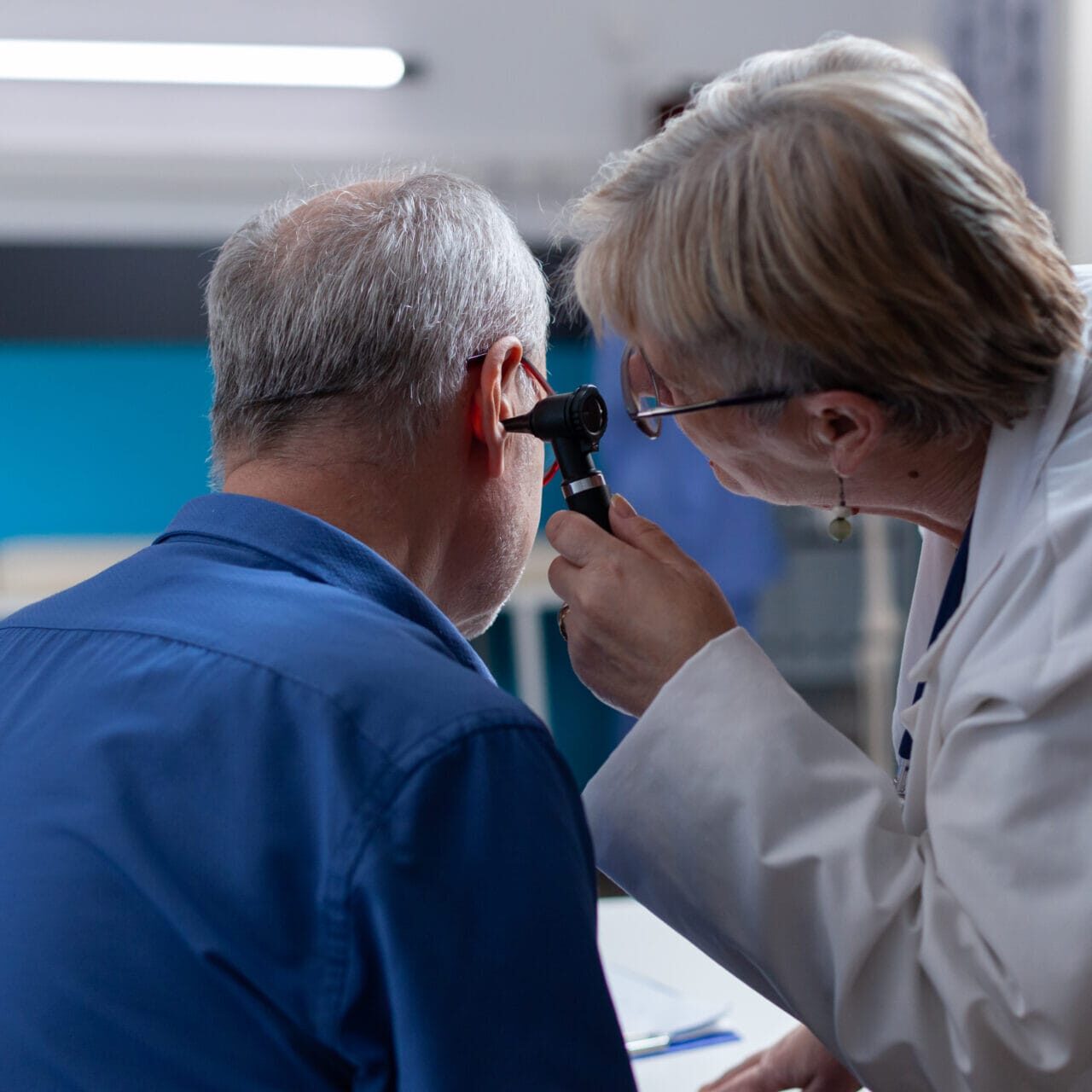 Physician holding otoscope to do ear consultation for patient at checkup visit. Woman doctor using otology instrument to examine infection and give medical advice to senior man.